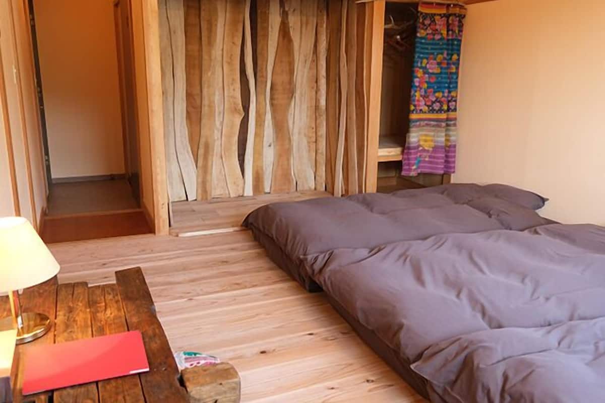 Um quarto com uma cama, um banco de madeira, uma pequena mesa e uma porta que dá para outro cômodo.