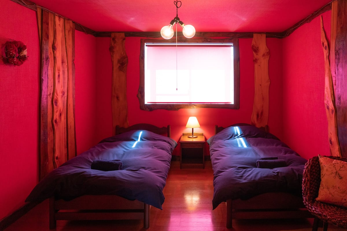 A room with two beds, a wooden floor, a window with a view, and a hanging light fixture.