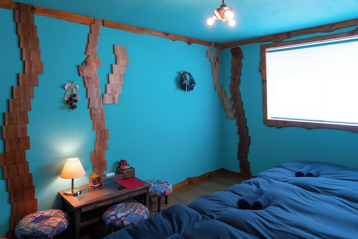A room with a blue wall, wooden wall decor, a bed with blue bedding, a wooden desk with a lamp, and a window with natural light.