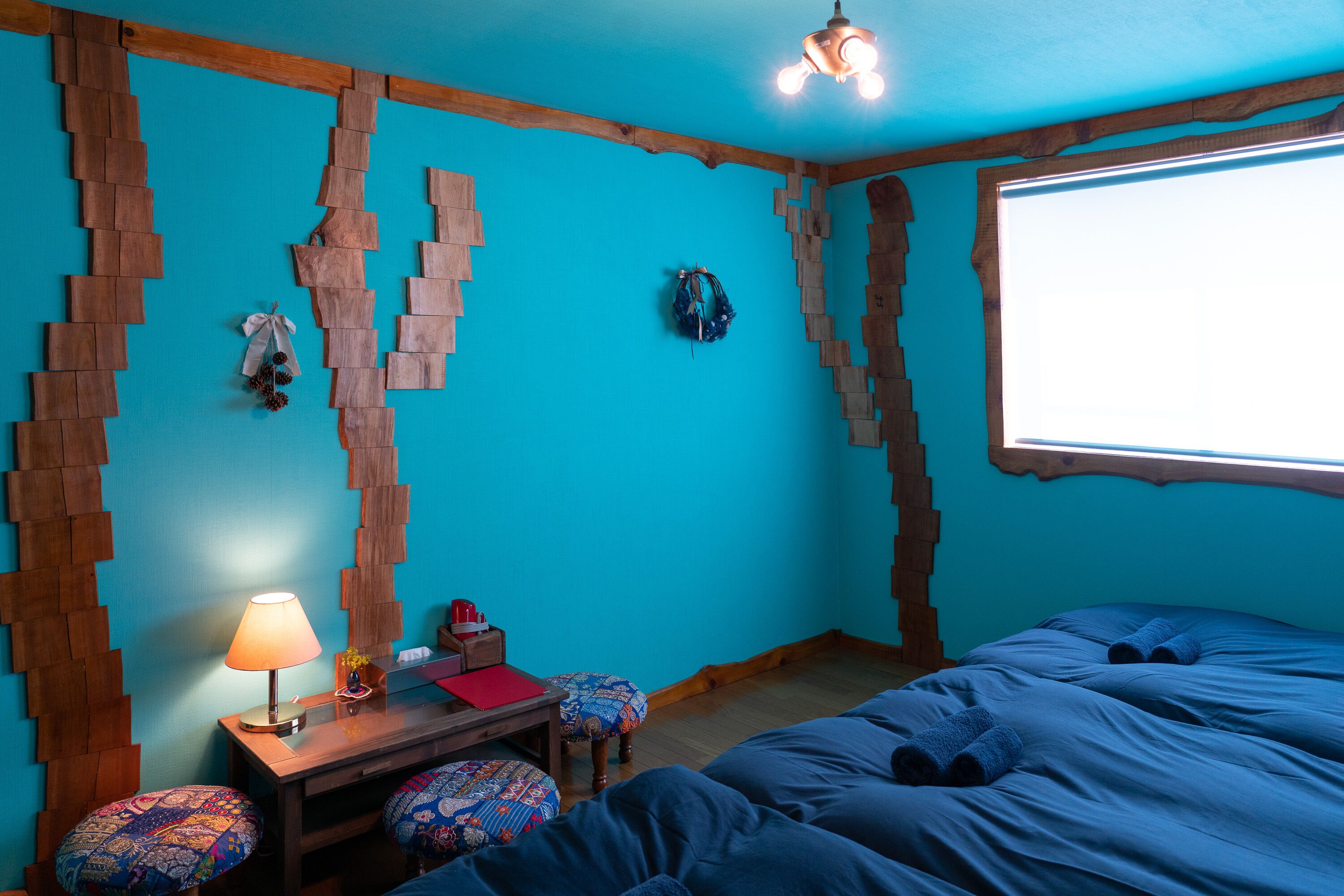 A room with a blue wall, wooden wall decor, a bed with blue bedding, a wooden desk with a lamp, and a window with natural light.