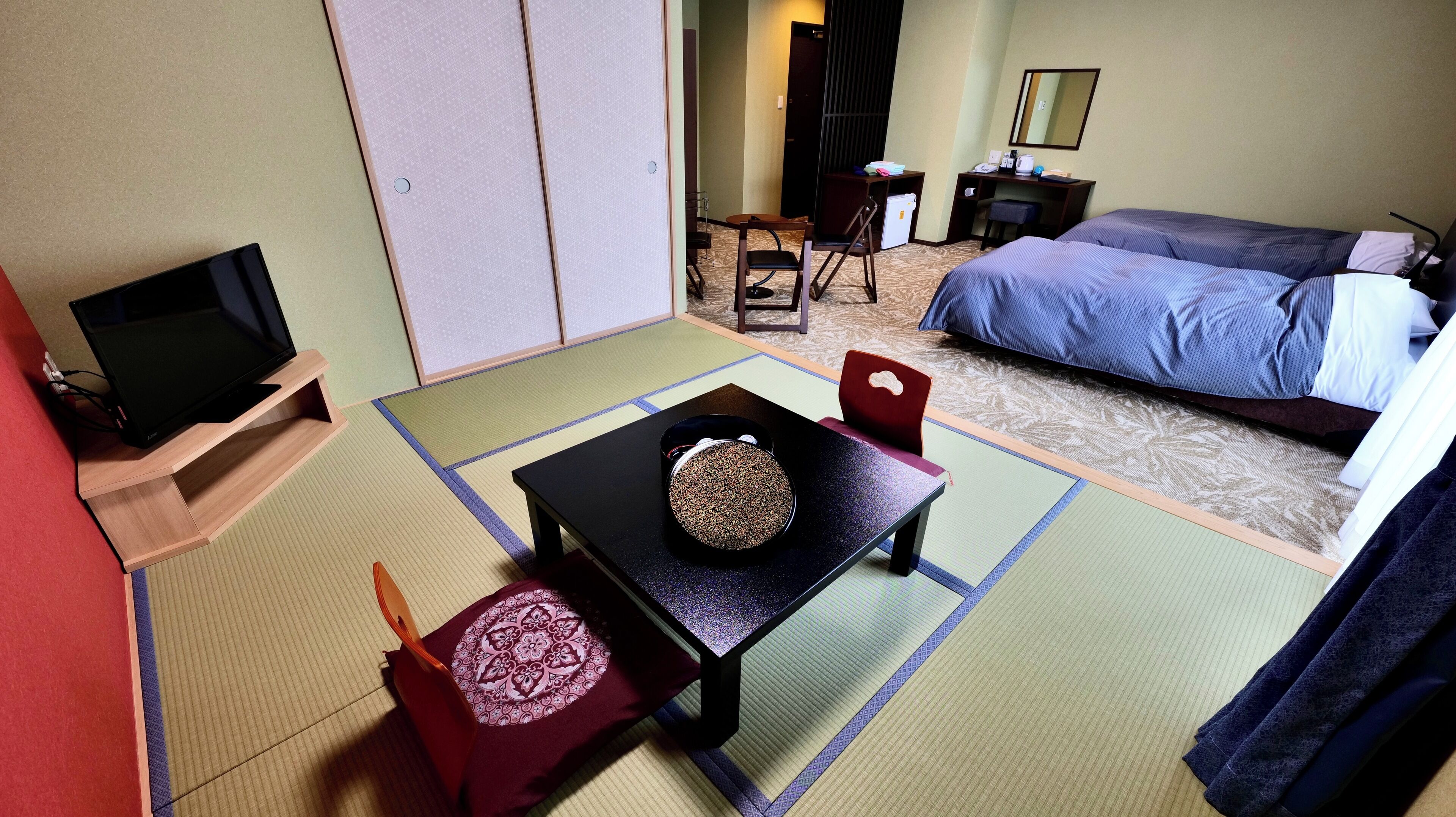 A hotel room with tatami flooring, a bed, a desk with a chair, a television, and a small table with a decorative item.