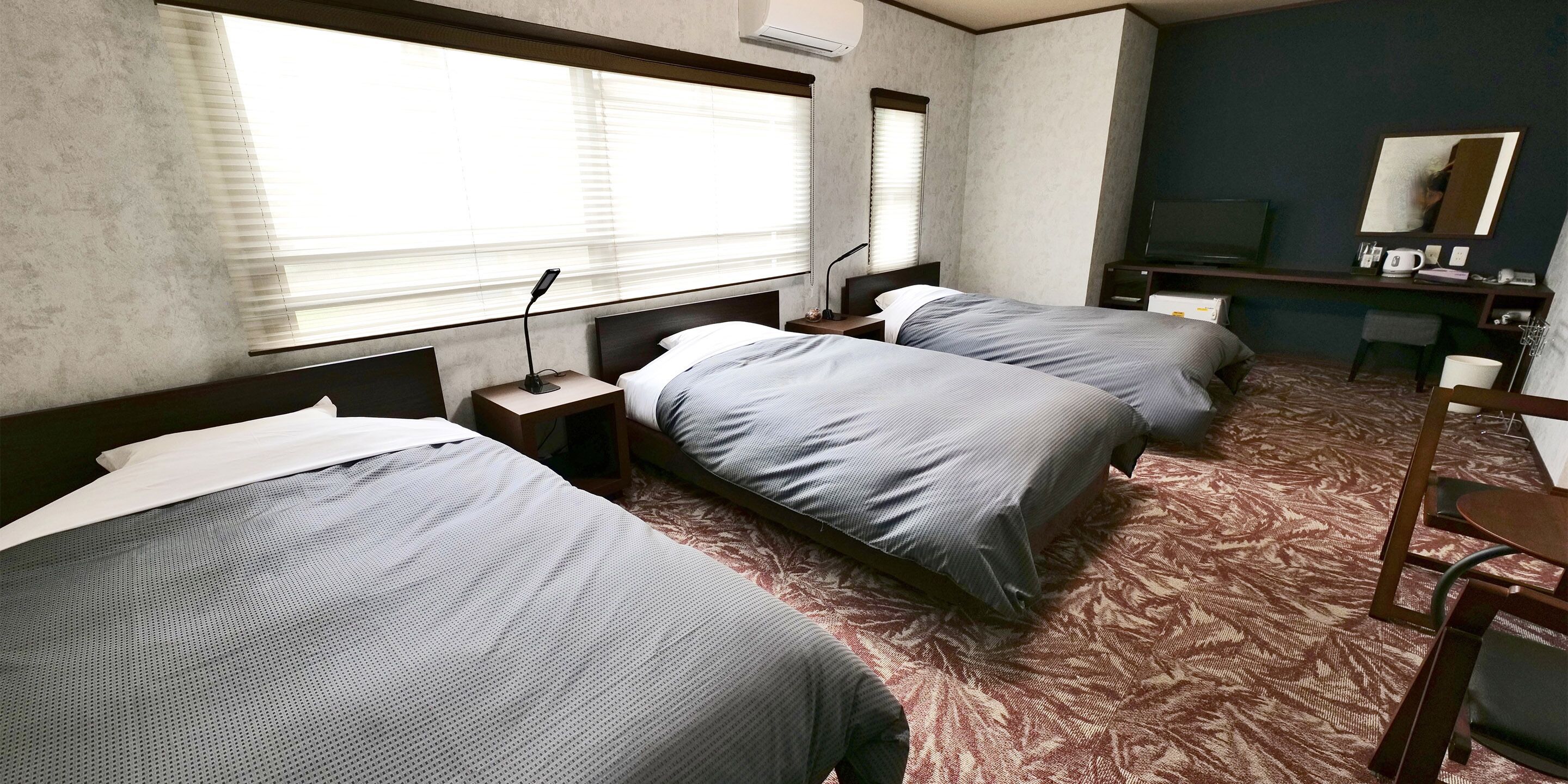 Habitación de hotel con dos camas, un escritorio, una silla, un televisor y un ventanal grande con persianas.