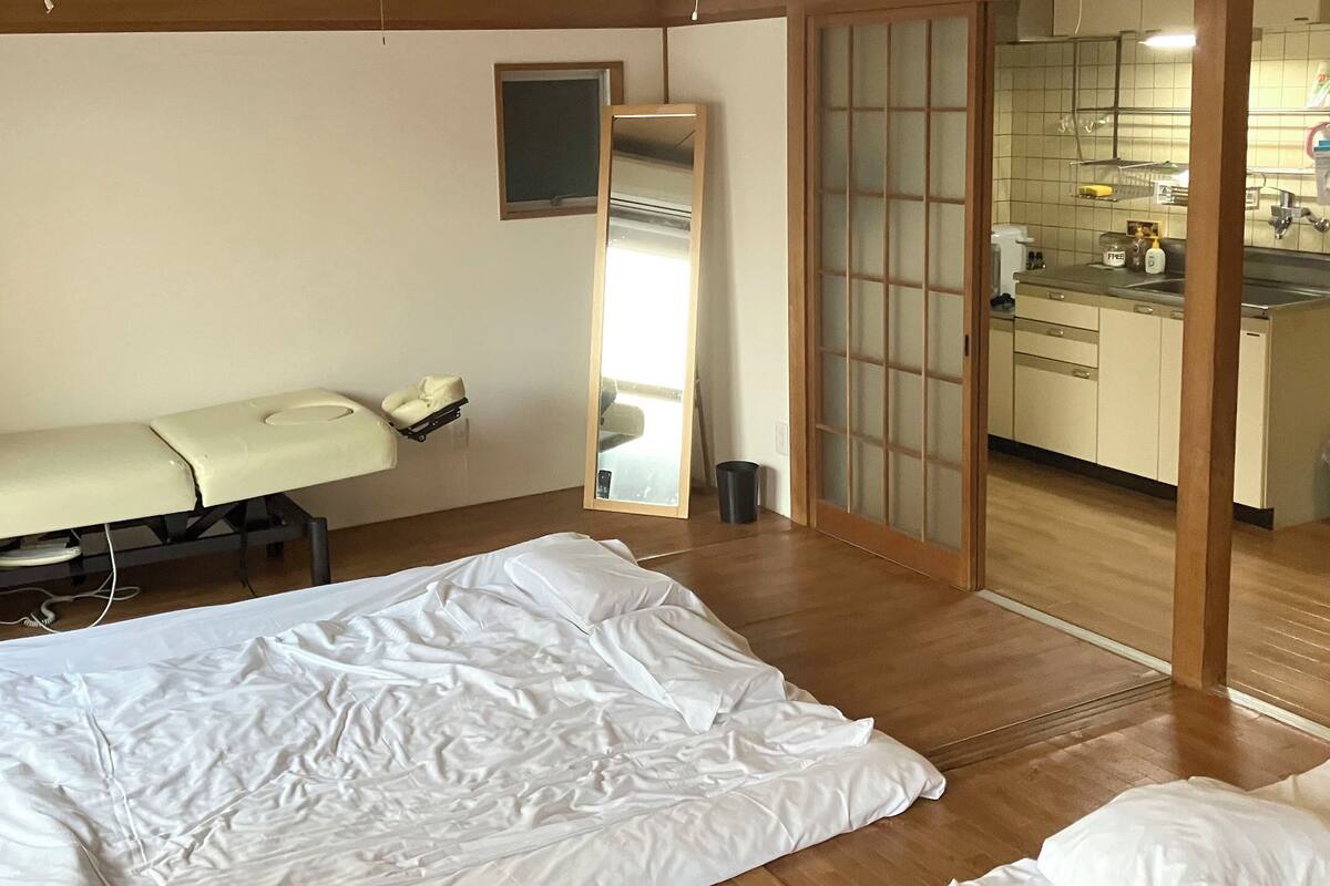 A room with a bed, a massage table, a wooden floor, a sliding shoji screen, and a bathroom with a sink and mirror.