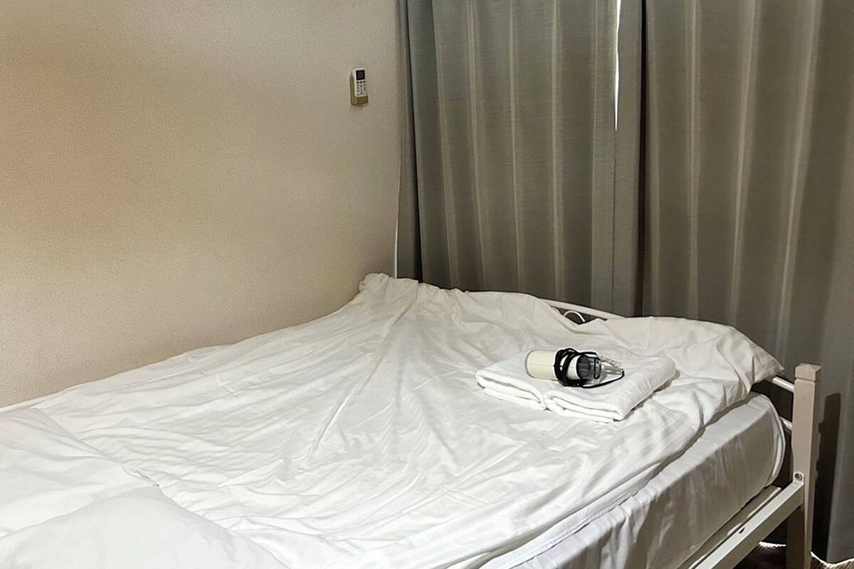 A single bed with white bedding, a bedside table with a bottle and glasses, and a wall-mounted phone.