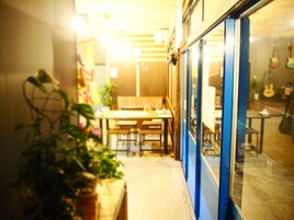 Un pasillo estrecho con un banco de madera y mesas, plantas en macetas y una pared azul con una ventana que refleja guitarras.