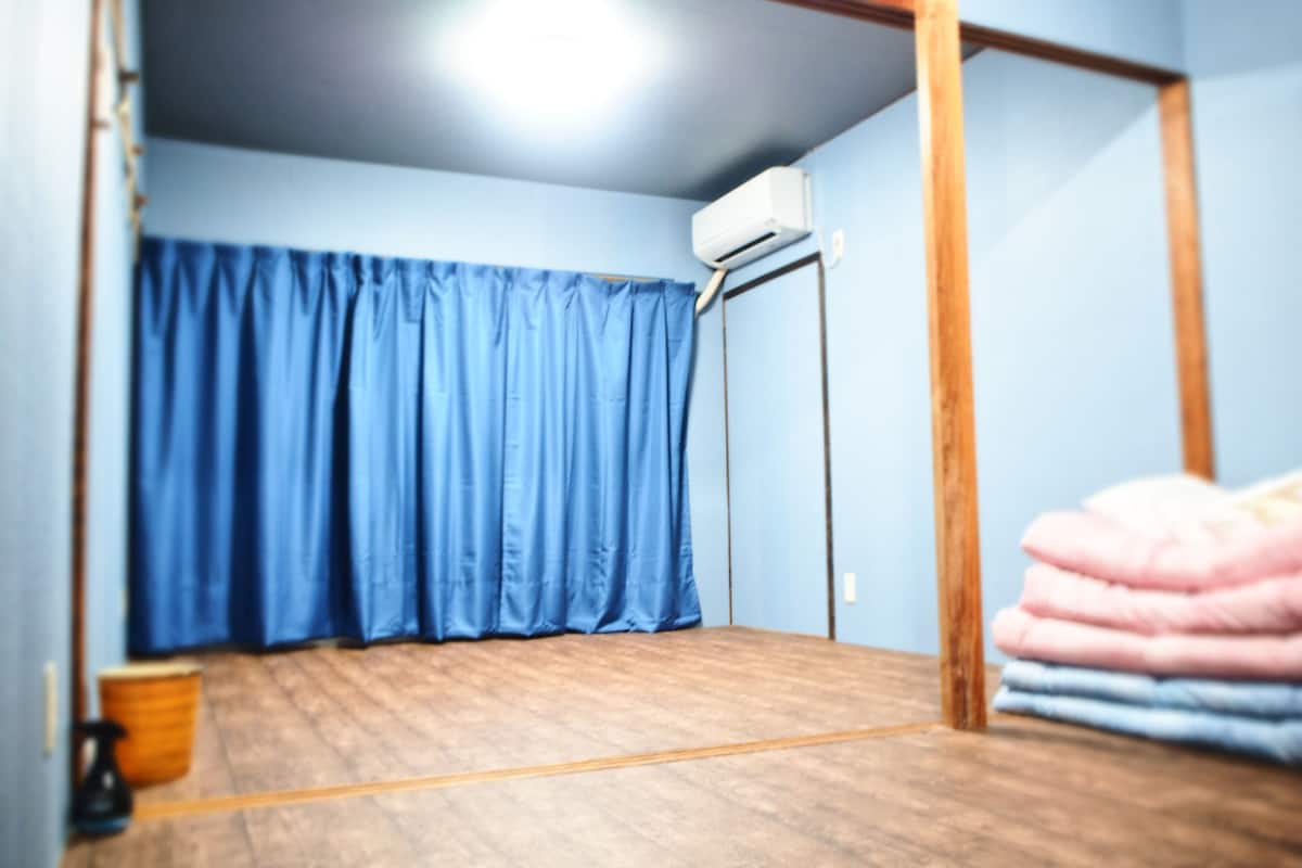 A room with a wooden floor, a blue curtain, and a stack of folded blankets.