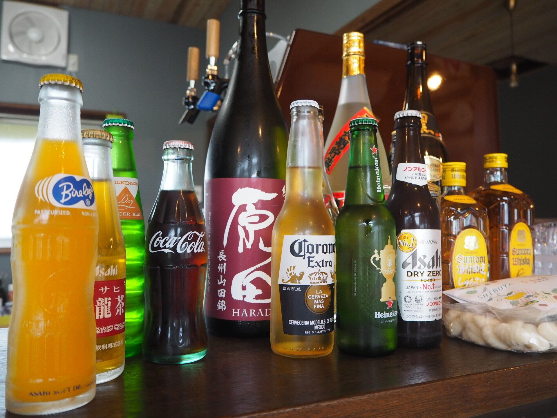 On voit une variété de bouteilles sur le comptoir d’un bar, notamment des marques comme Coca-Cola, Corona et Heineken.