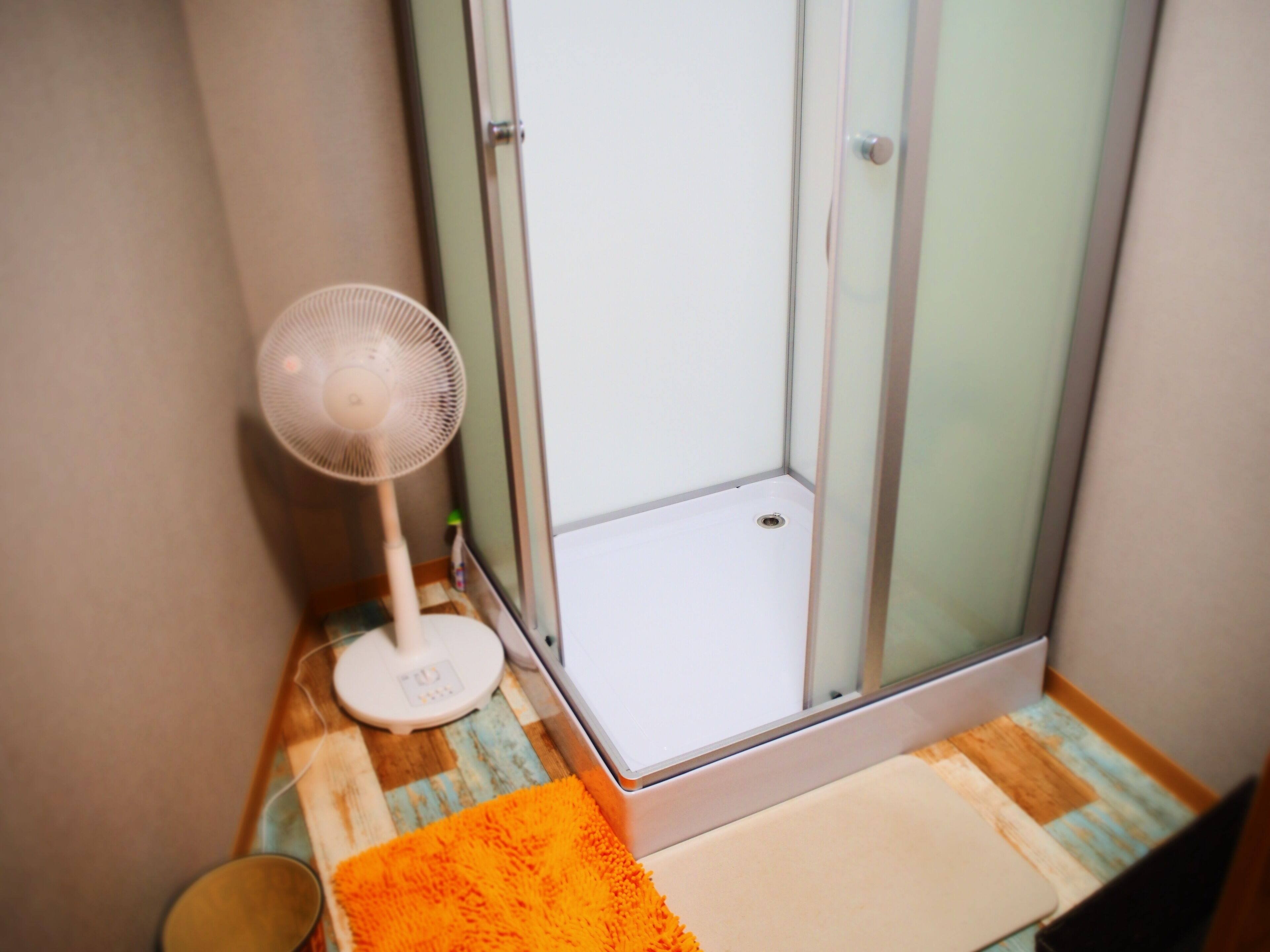 A bathroom with a shower, a white fan, and a wooden floor.