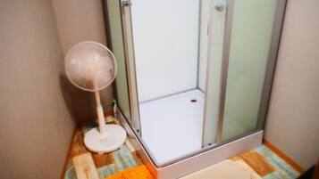 A bathroom with a shower, a white fan, and a wooden floor.