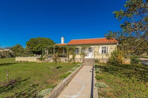 A two-story house with a red-tiled roof, a stone pathway leading to the entrance, and a garden with various plants and trees. - Holiday house TRSJA with swimming pool (Linardići)