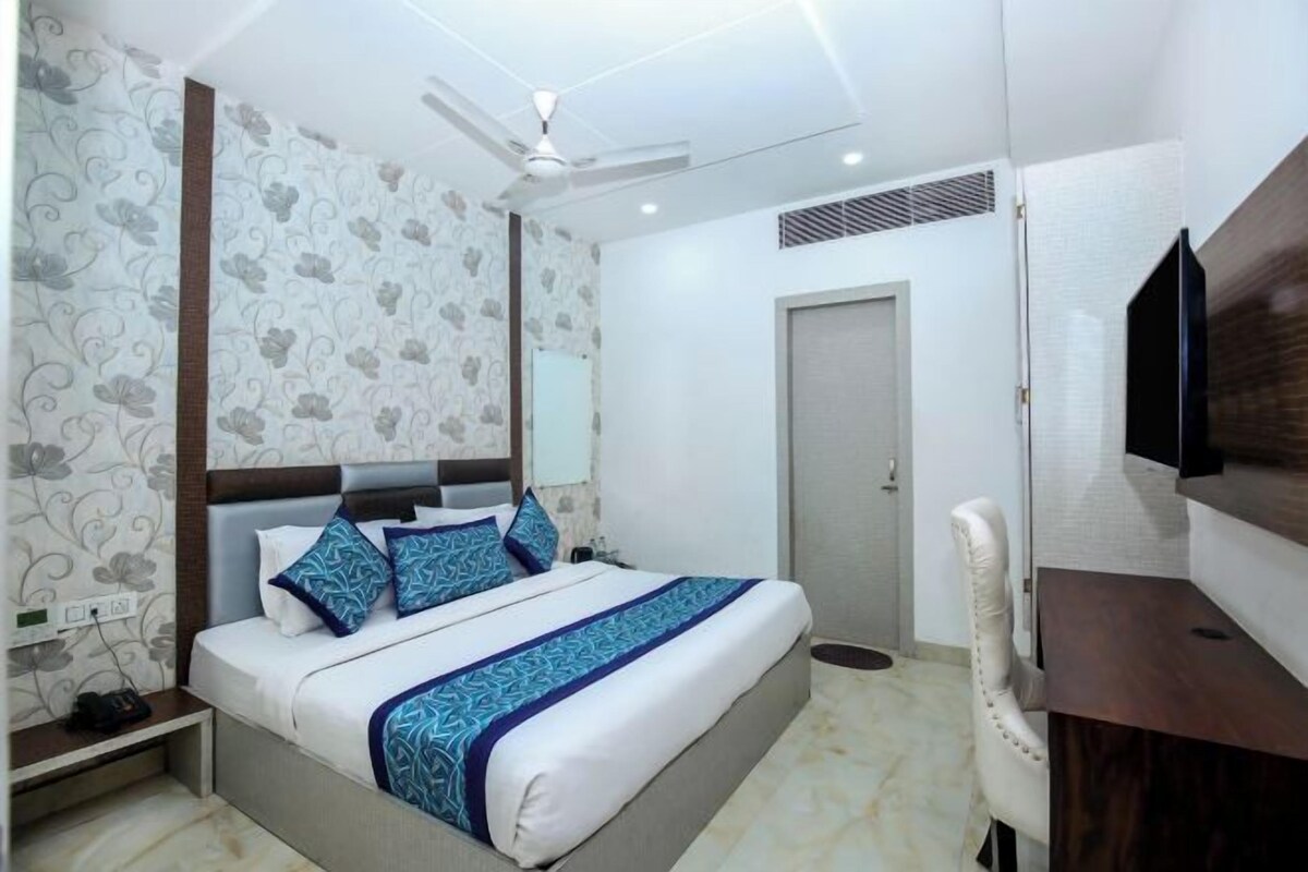A hotel room with a bed, a desk, a chair, a TV, a ceiling fan, and a door.