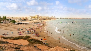 Una playa con gente tomando el sol y nadando, una orilla arenosa y edificios al fondo.