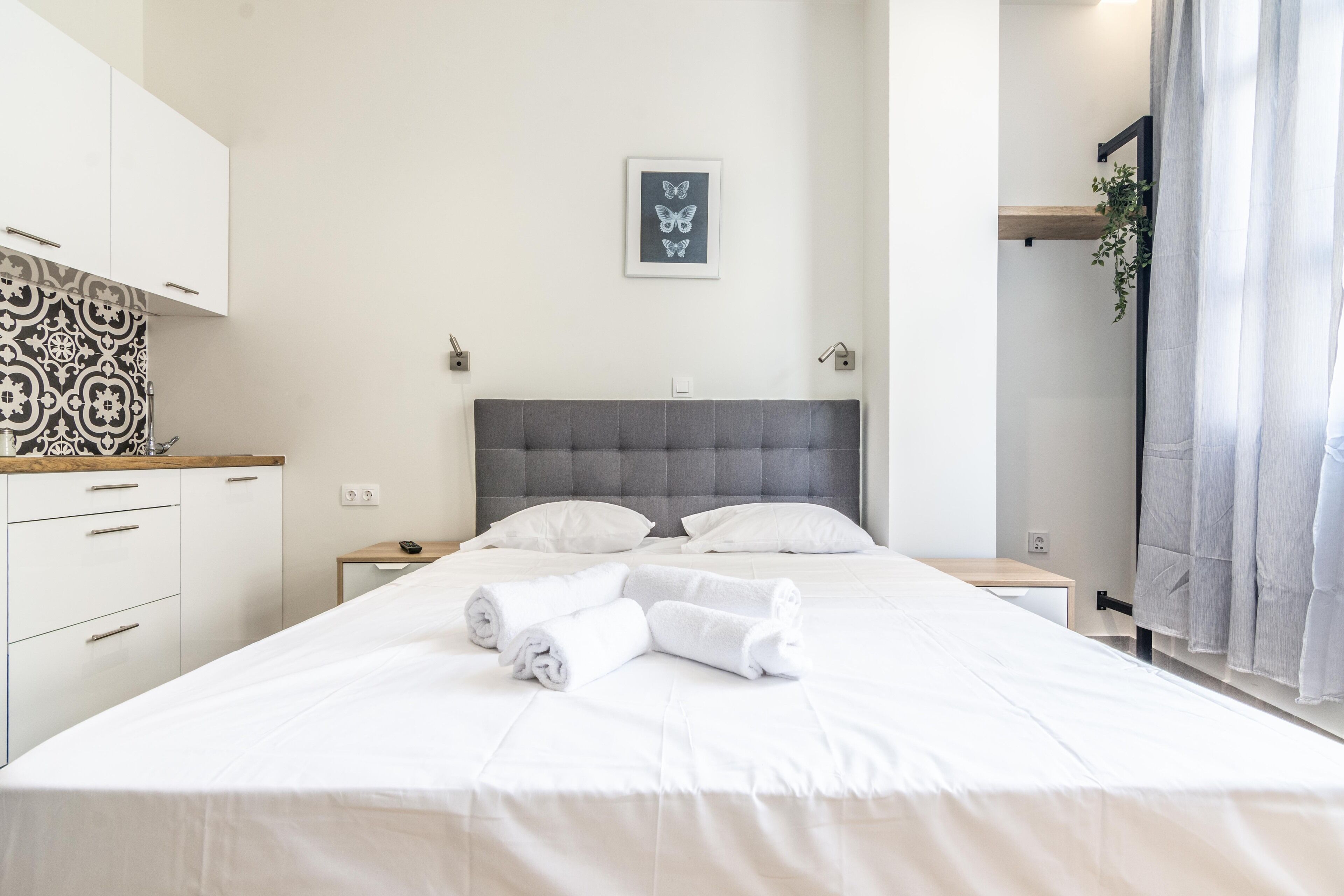 Un lit bien fait, avec du linge de lit blanc et des serviettes pliées, une tête de lit rembourrée de couleur grise, un interrupteur fixé au mur et un tableau représentant un papillon.