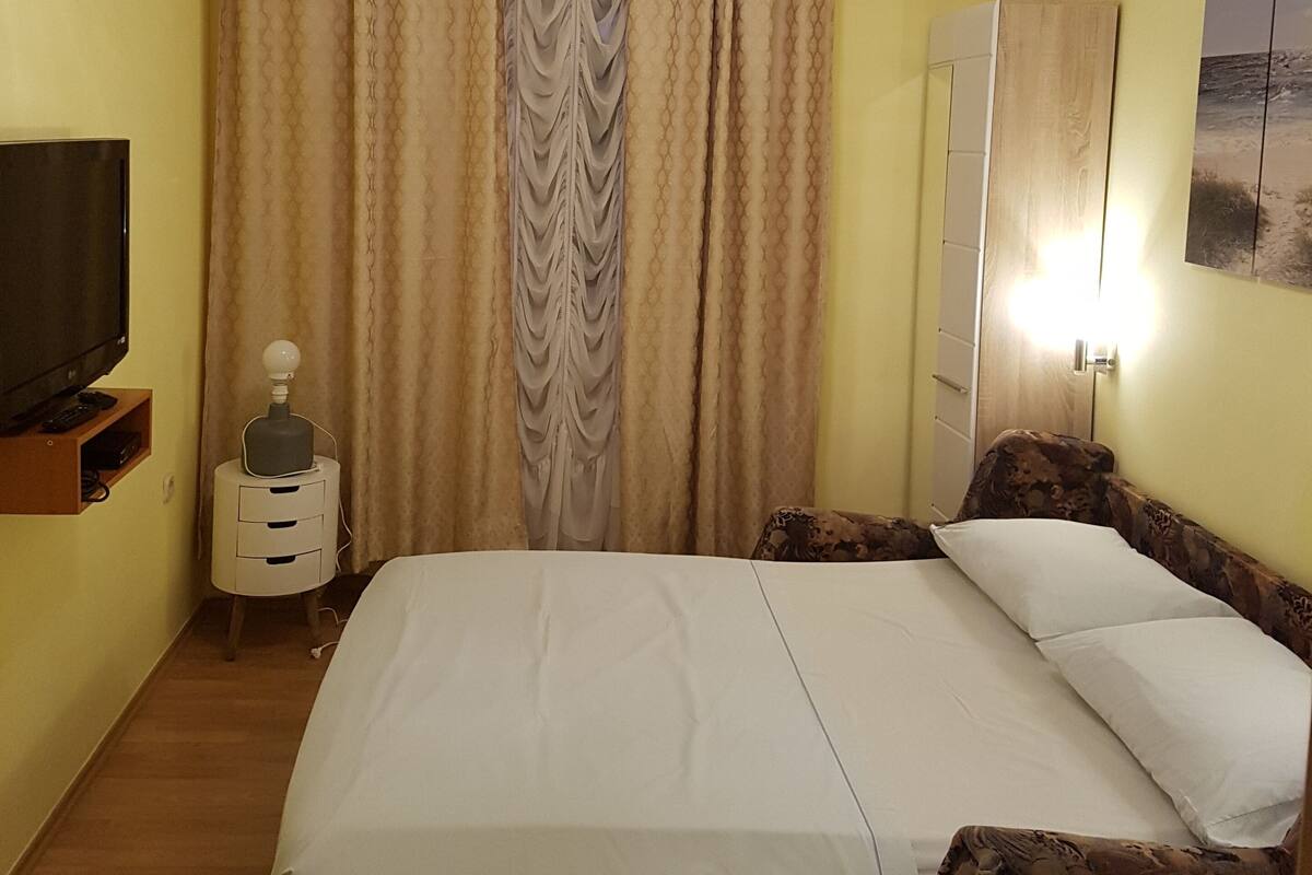 A hotel room with a bed, curtains, a television, a small table with a lamp, and a wardrobe.