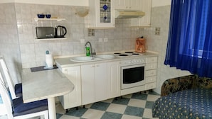A compact kitchen with white cabinets, a stove, a sink, and a microwave. There's a chair and a patterned sofa. - Apartments Aurora (Zadar)