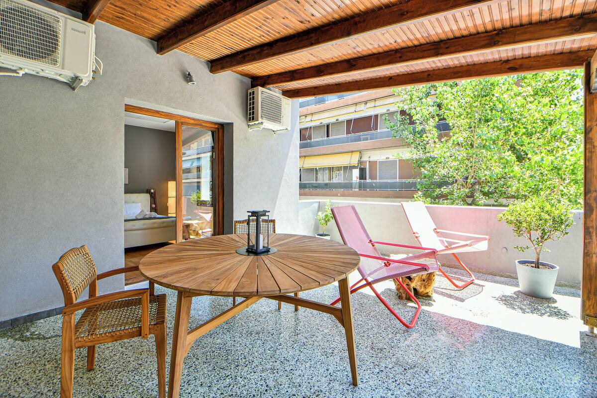 Eine überdachte Terrasse im Freien mit einem runden Holztisch und Stühlen, einem Blumentopf und Blick auf ein Gebäude im Hintergrund.