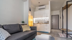 A modern living room with a grey sofa, a small white table, and a kitchen area with white cabinets and a patterned backsplash. - Plenty Apartments Zeus (Athens)