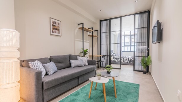 Een moderne woonkamer met een grijze bank, een witte salontafel en een groen vloerkleed. Er is een groot raam met gordijnen, een aan de muur bevestigde televisie en een plant op de salontafel.