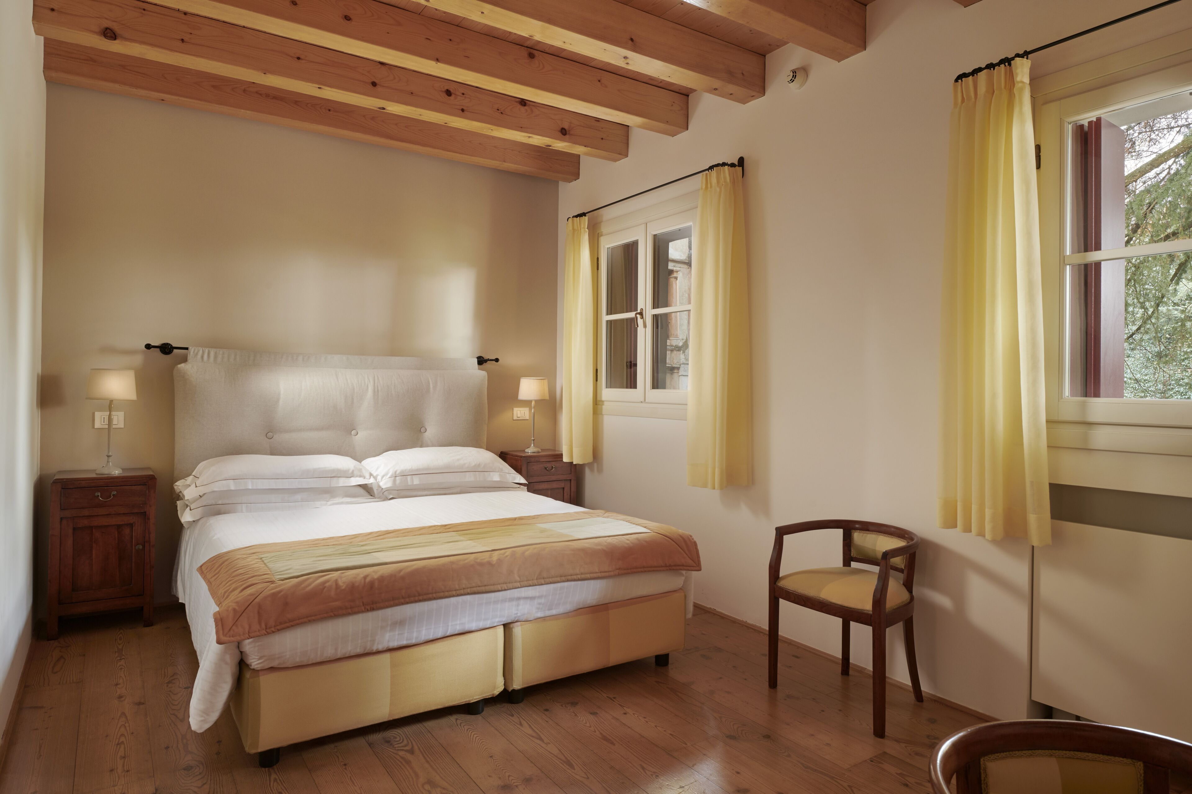A bedroom with a large bed, wooden floor, two bedside tables with lamps, a chair, and a window with curtains.
