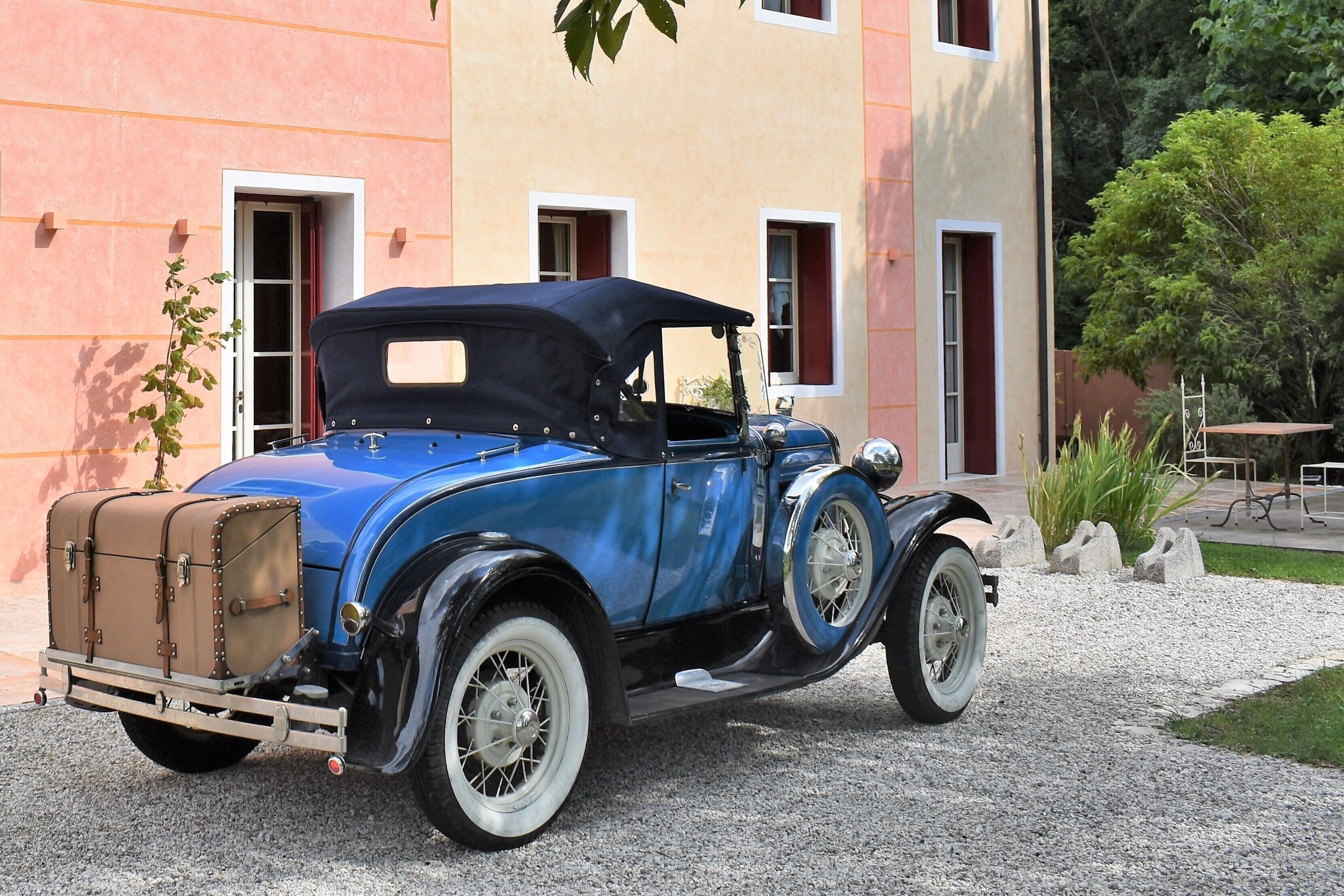 Un coche azul antiguo aparcado en un camino de grava frente a un edificio con ventanas de marco rojo y fachada color beige.