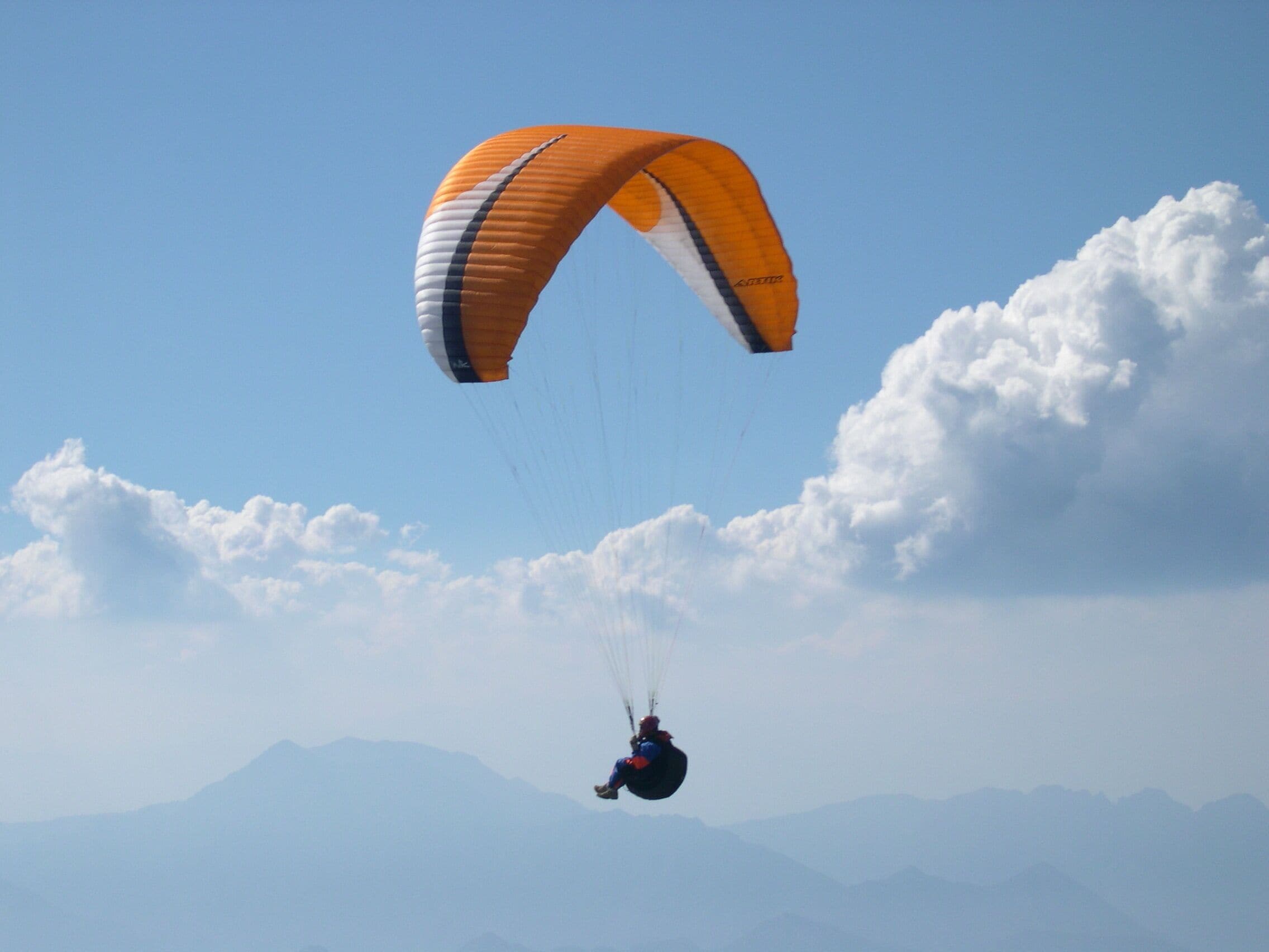 A paraglider with an orange canopy flying over a mountainous landscape.