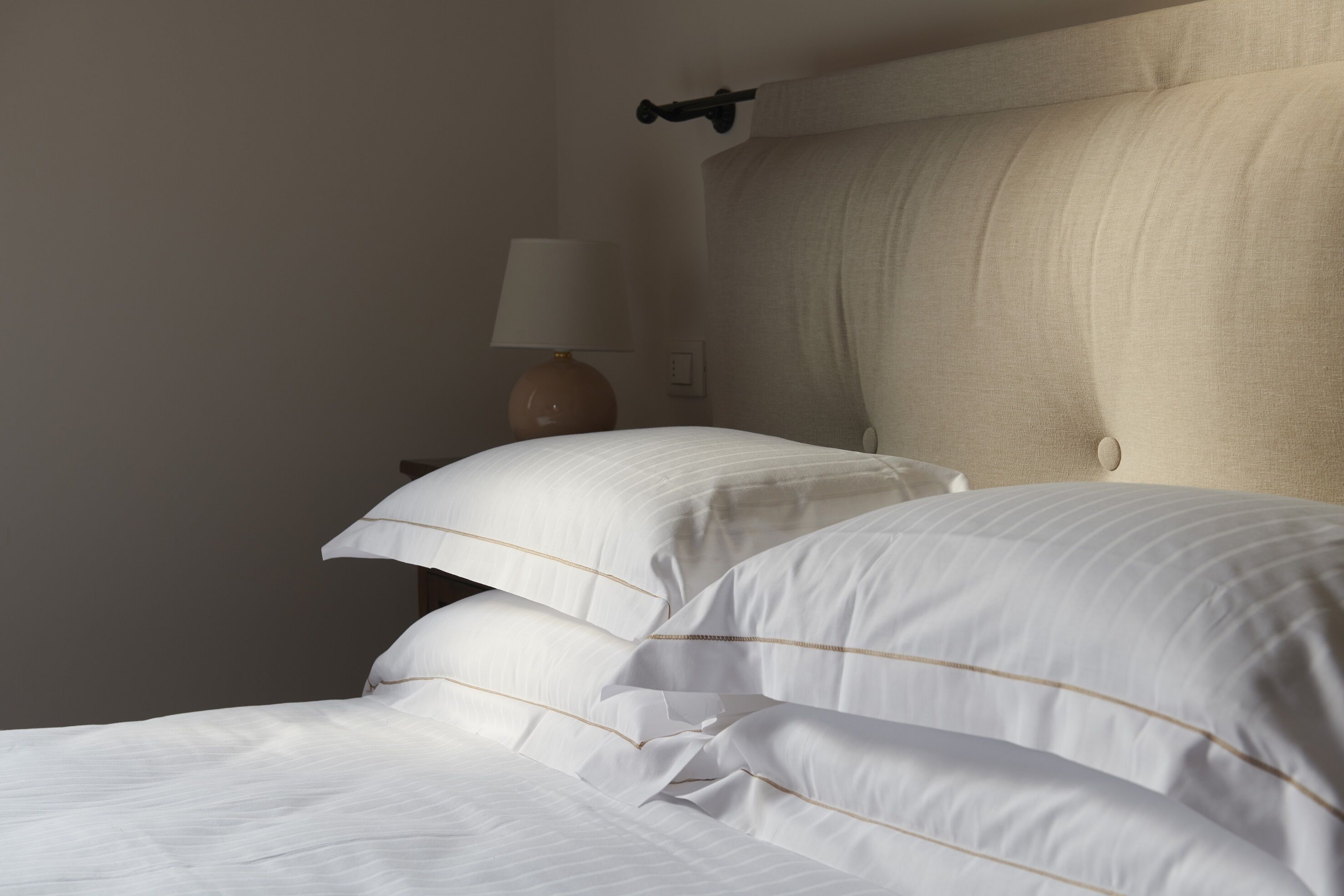 Una cama bien tendida con sábanas blancas y detalles dorados, una lámpara de noche y un interruptor de luz en la pared.