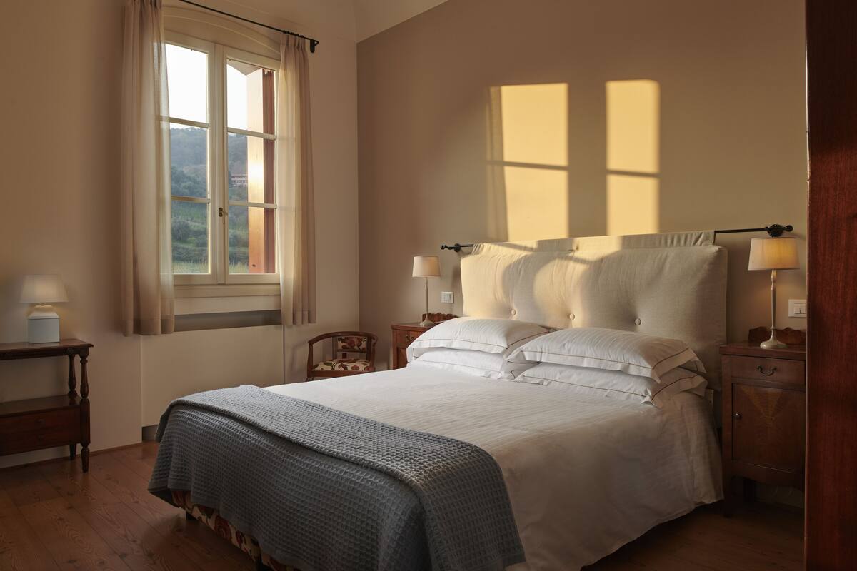 Ein Schlafzimmer mit einem großen Bett, zwei Nachttischen, einem Beistelltisch, einem Fenster mit Vorhängen und einer Lampe auf jedem Nachttisch.