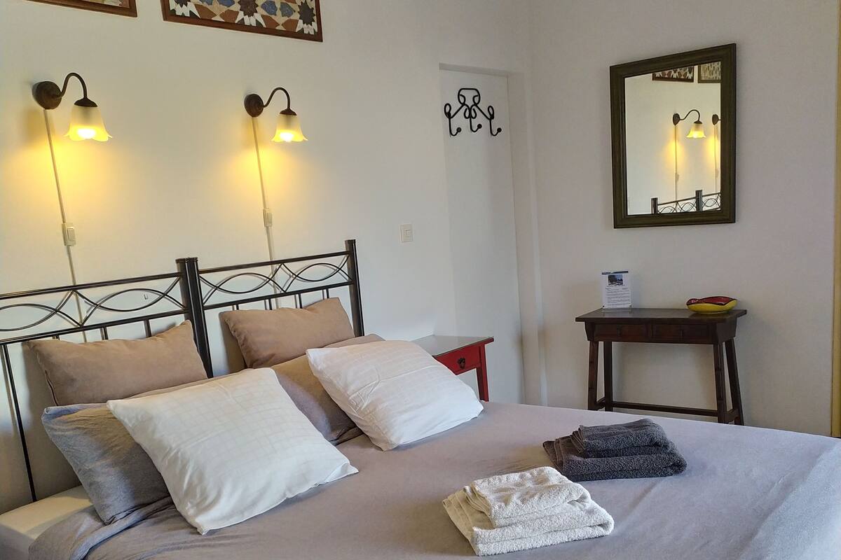 A neatly made bed with a grey bedspread, two pillows, and a folded towel. There are wall-mounted lights, a small table with a bowl, and a mirror.