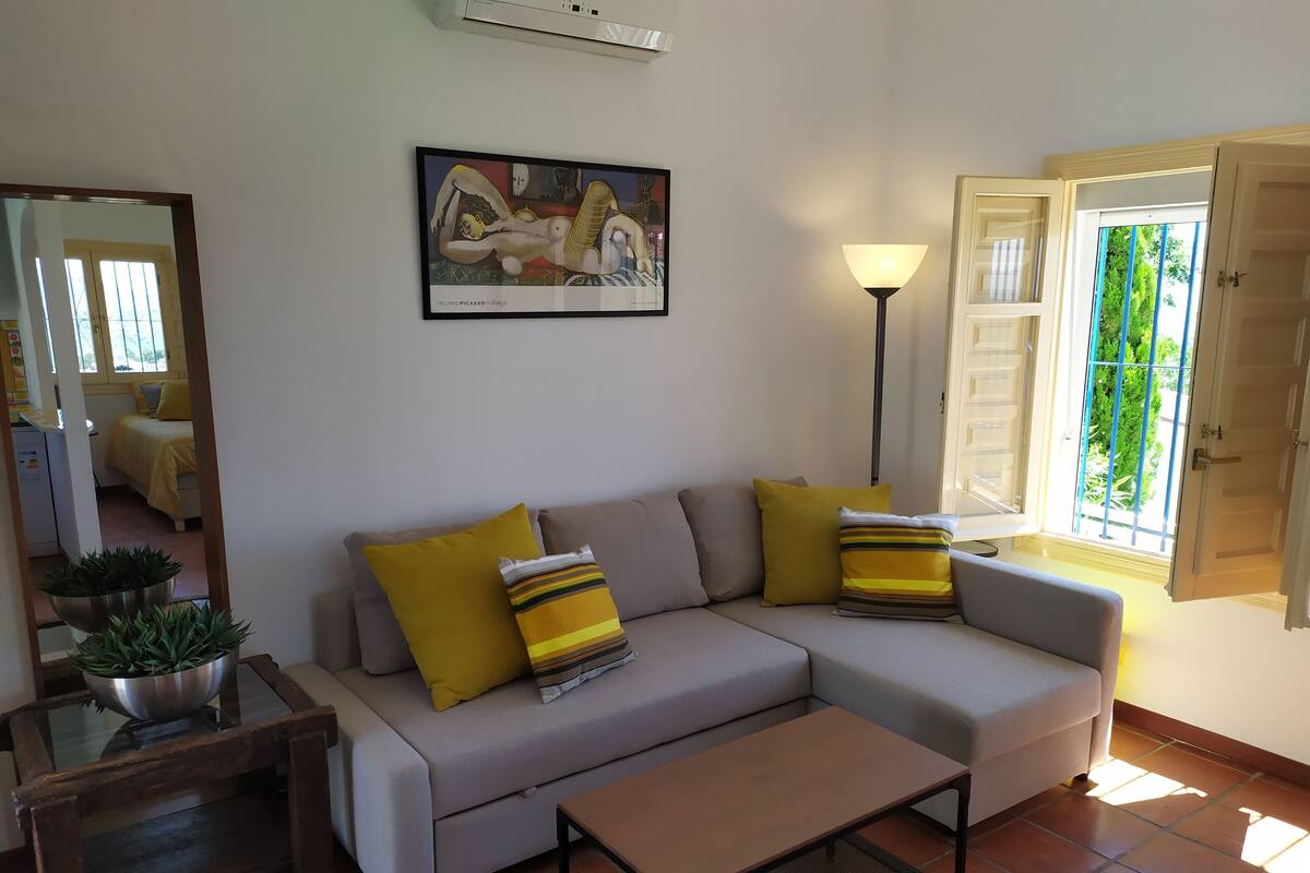 A living room with a sofa, a coffee table, a floor lamp, a painting, and a window with a view of greenery.