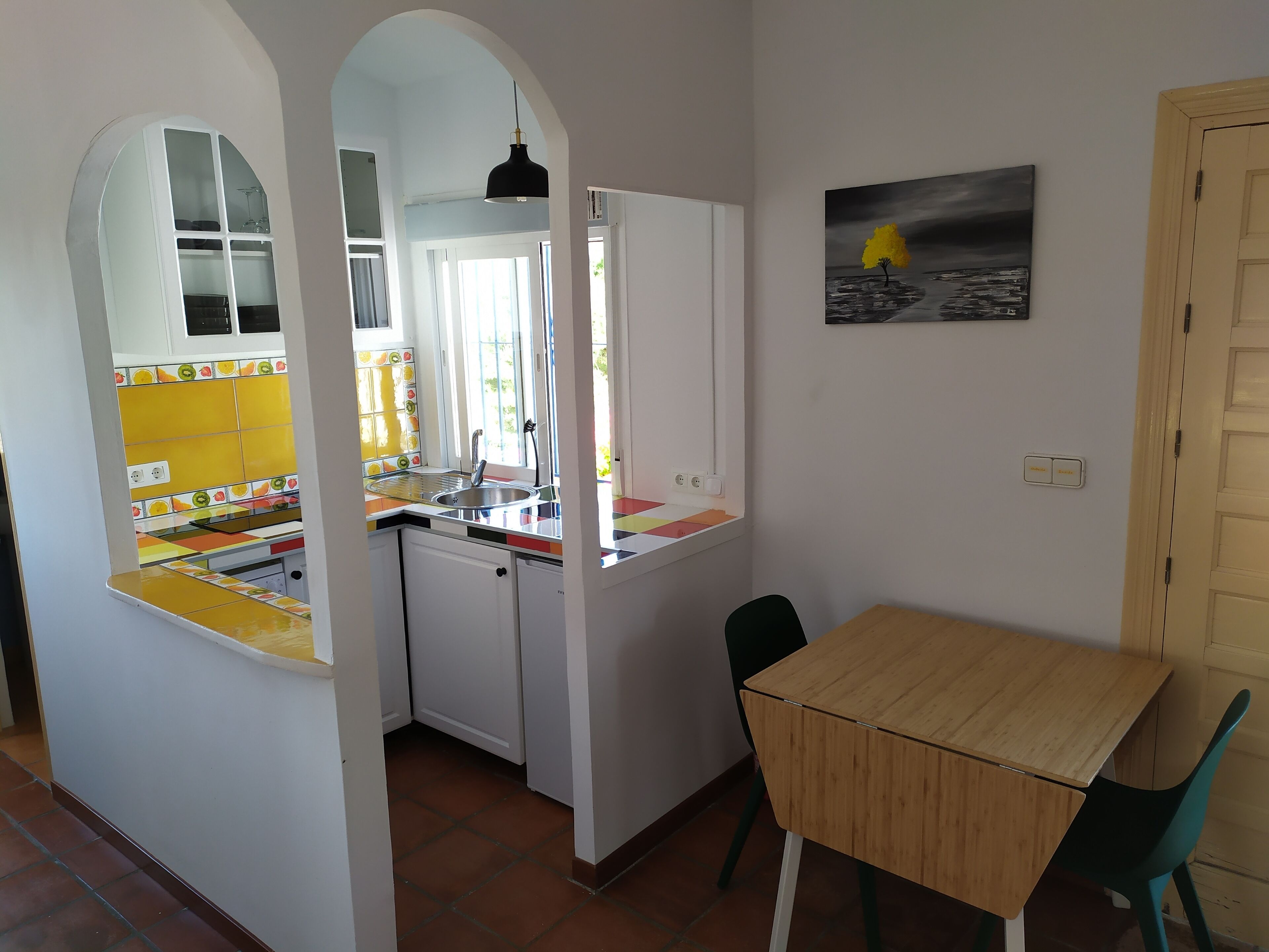 A compact kitchen with a window, a dining area with a table and chairs, and a painting on the wall.