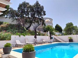 Una piscina con sillones de descanso, una planta en maceta y un edificio de fachada blanca y ventanas arqueadas al fondo.