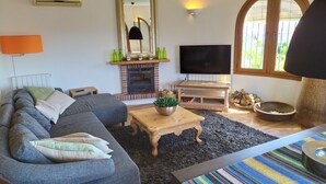 A living room with a sofa, a fireplace, a television, and a window with blinds. - Bed and Breakfast La Madrugada (Còmpeta)