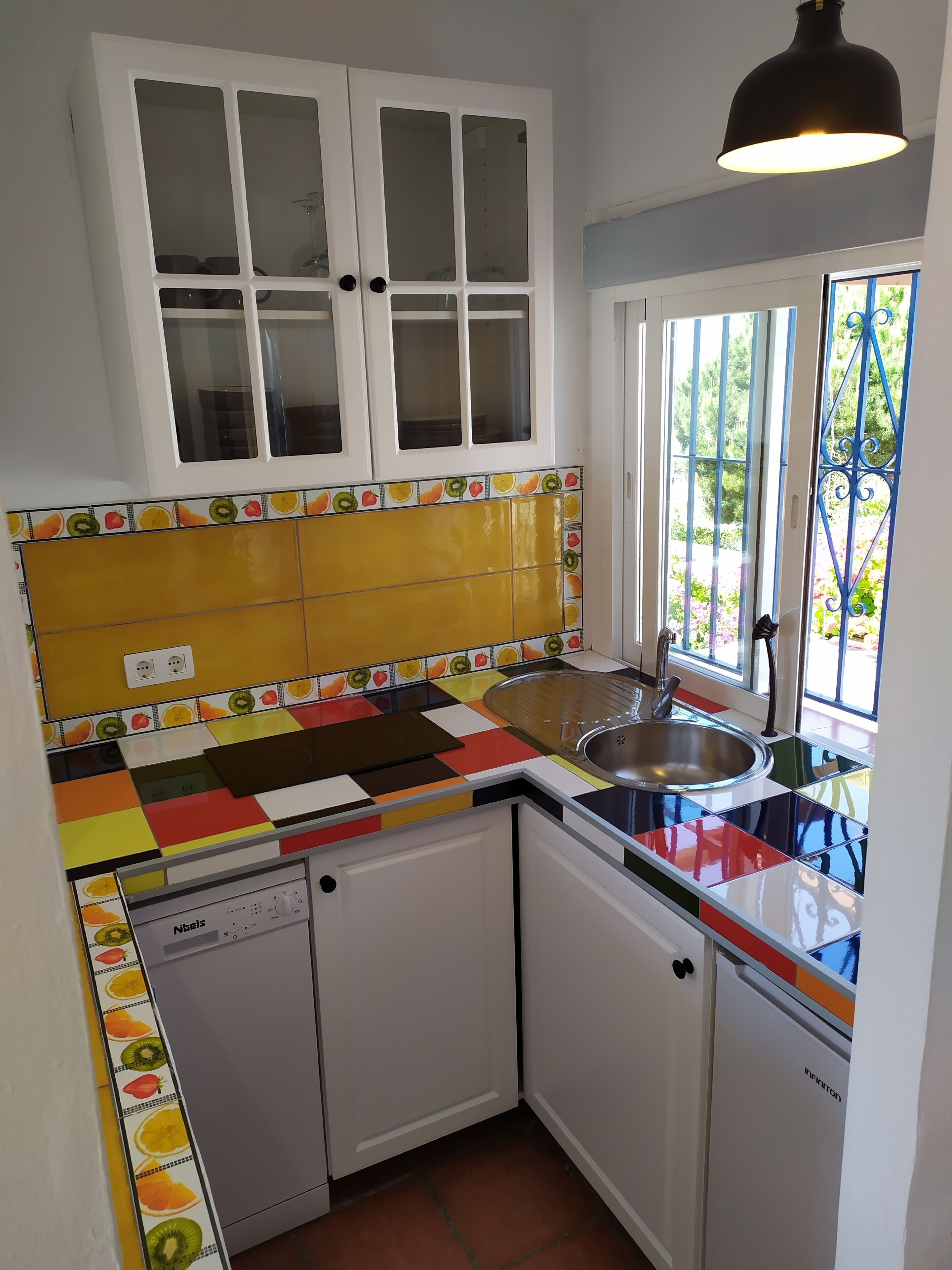 A compact kitchen with white cabinets, a colorful backsplash, and a window with a view of greenery.