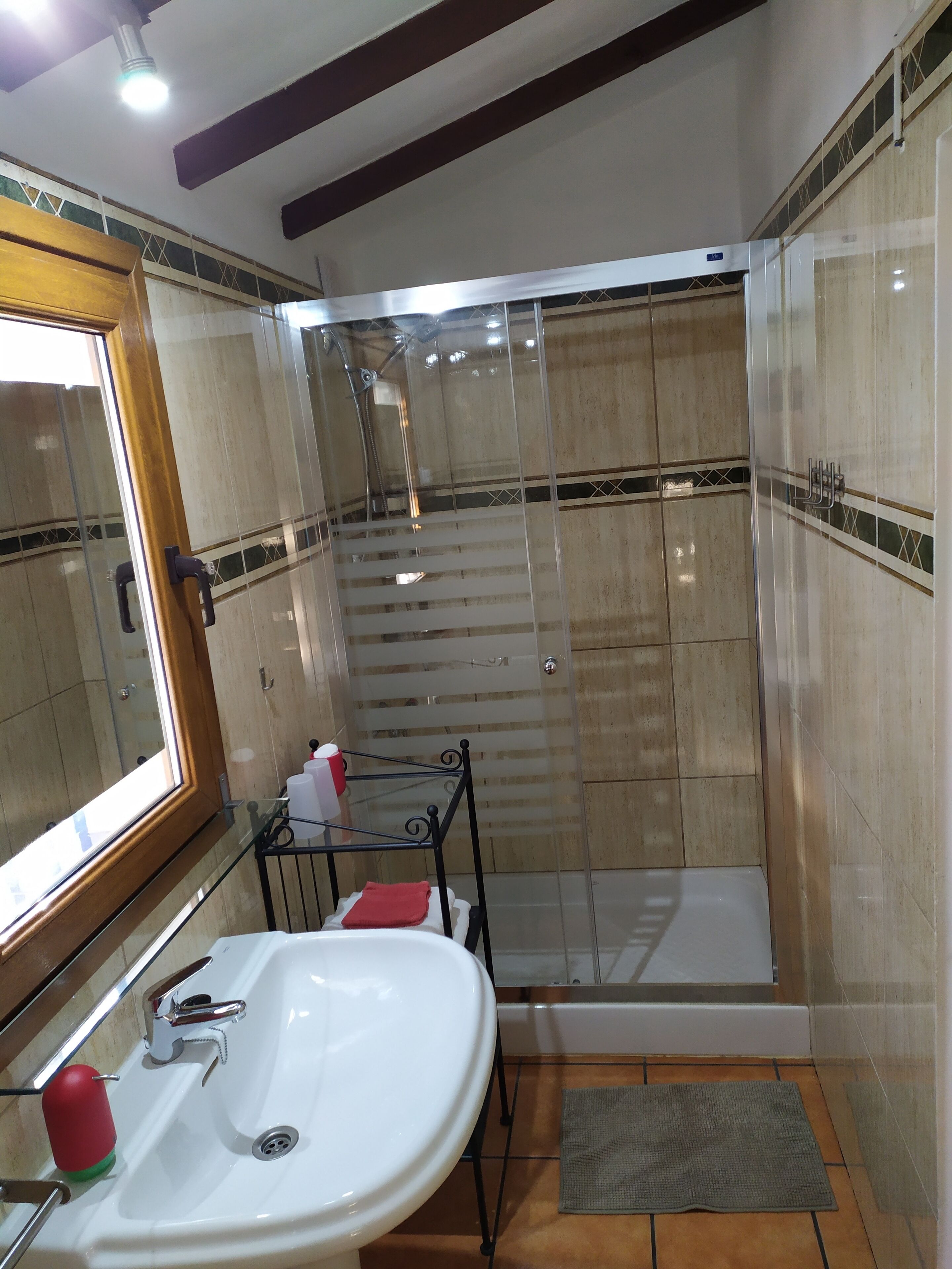 A bathroom with a glass-enclosed shower, a sink with a red soap dispenser, and a mirror with a wooden frame.