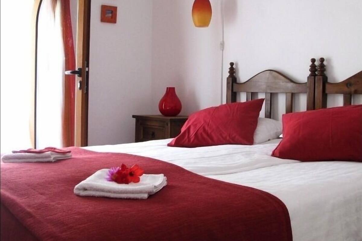A neatly made bed with red and white bedding, a wooden headboard, and a red vase on a nightstand.