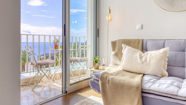 A balcony with a view of the sea, a sofa with cushions, a floor lamp, and a small table with a plant. - Elsas Place, a Home in Madeira (Funchal)