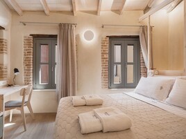 Un dormitorio con un escritorio de madera, una silla, una cama con cabecera acolchada y dos ventanas con cortinas.