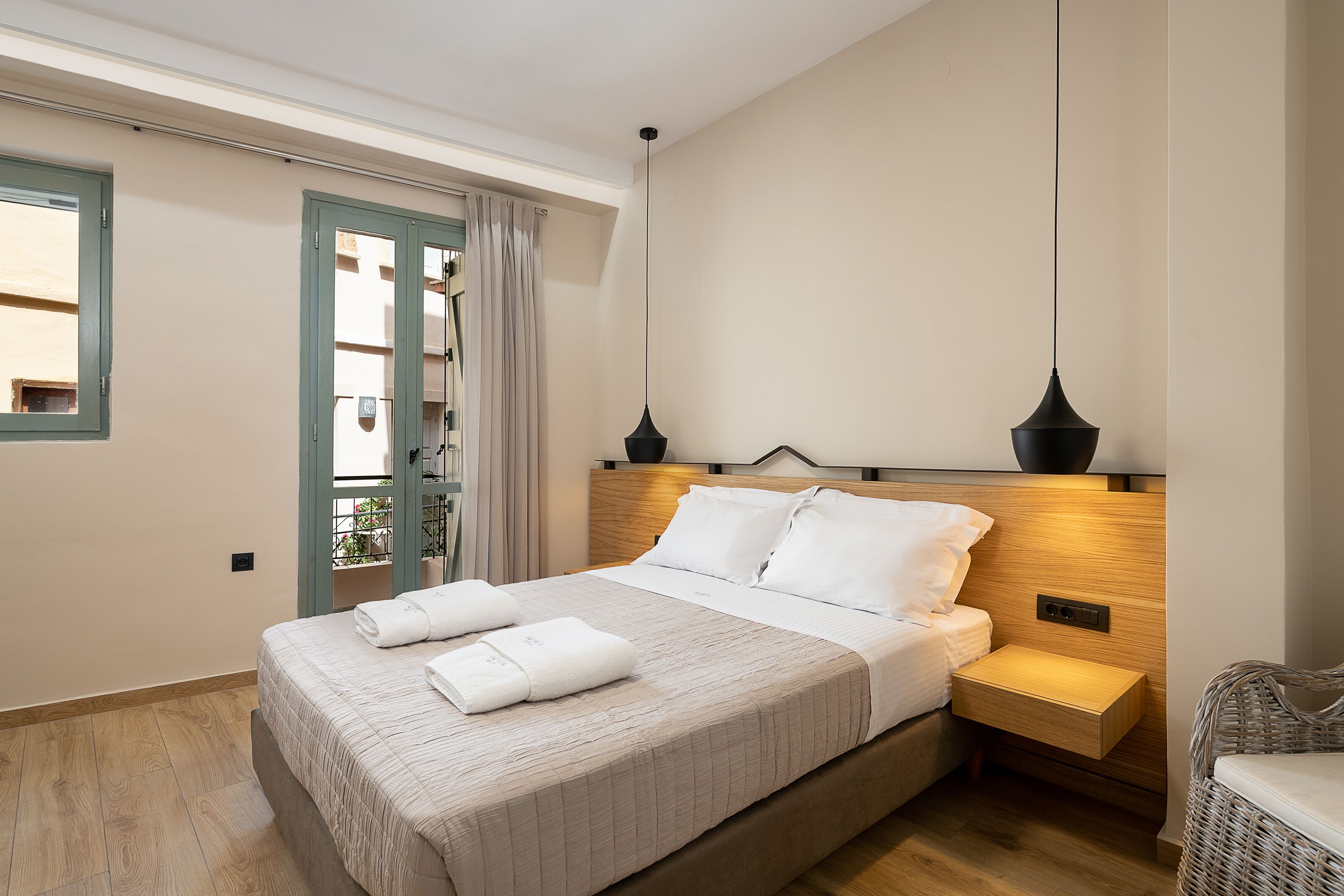 Een net opgemaakt bed met witte lakens en twee kussens, een nachtkastje met een lamp, en uitzicht op een balkon met een balustrade.