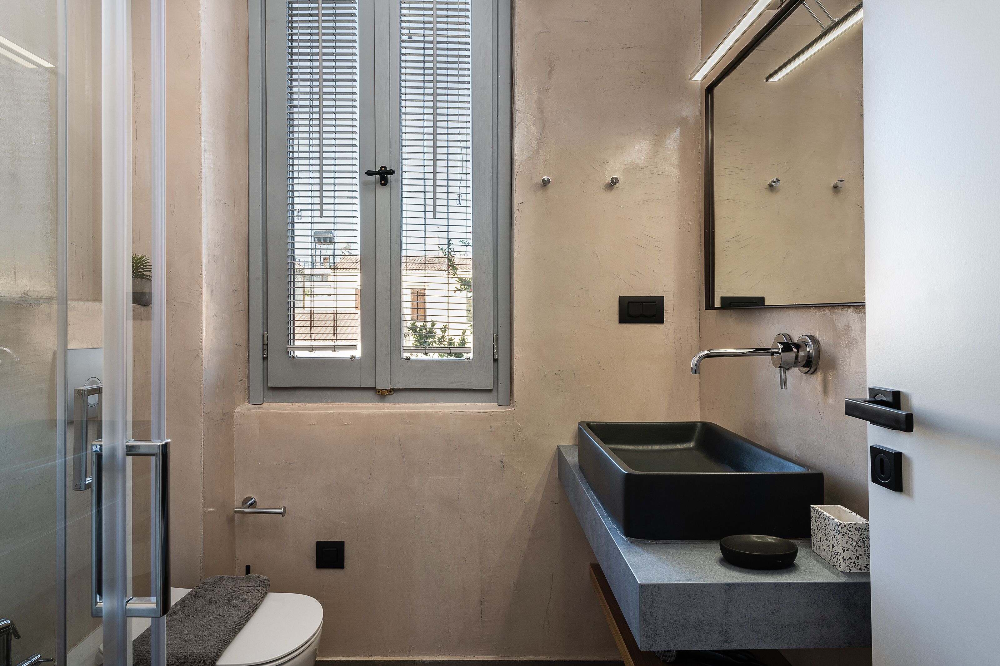 A modern bathroom with a glass shower enclosure, a toilet, a sink with a black basin, and a window with blinds.