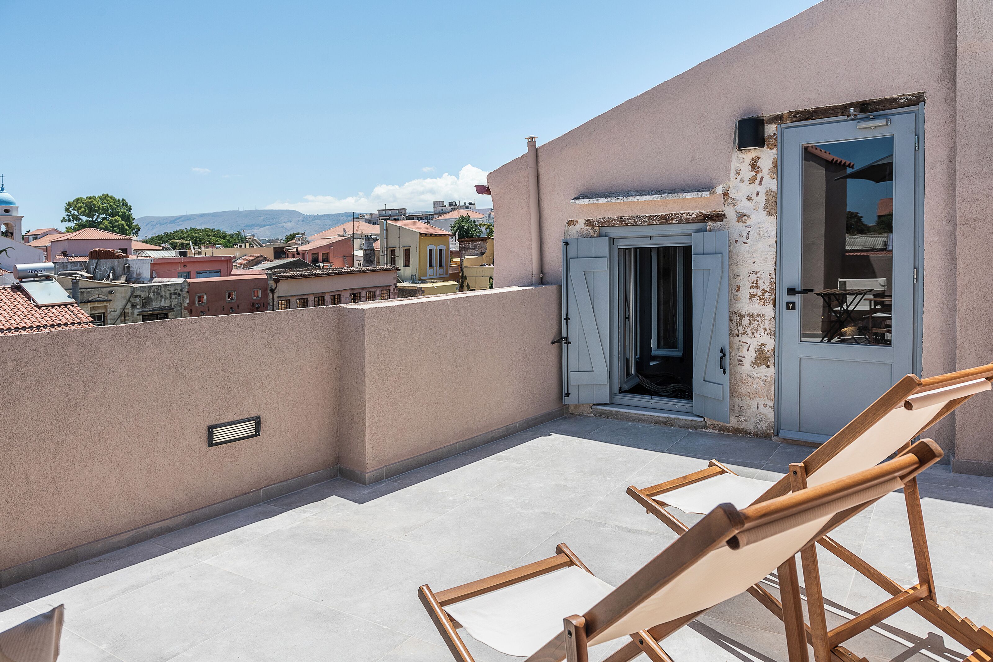 A rooftop terrace with a view of the city, a door leading to an interior space, and two wooden chairs.