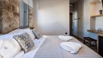 A hotel room with a bed, two pillows, a bedside table, a wooden stool, and a wall with a stone texture.
