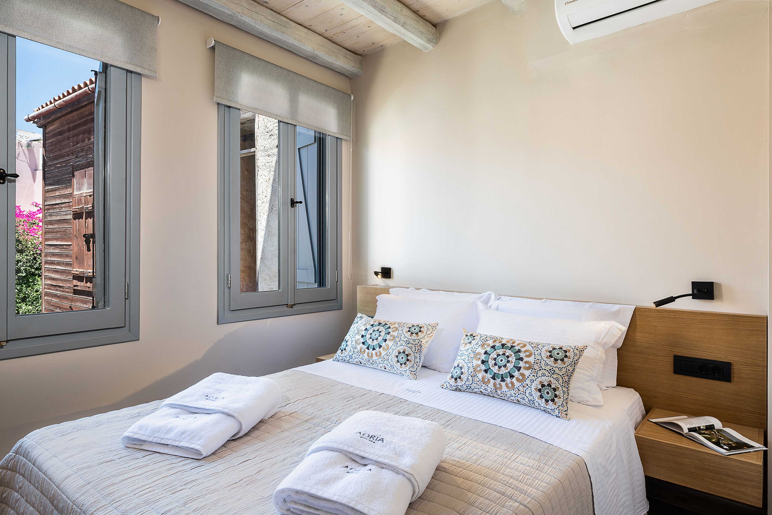 A neatly made bed with white linens and decorative pillows, a bedside table with a magazine, and a window with a view of a building and pink flowers.
