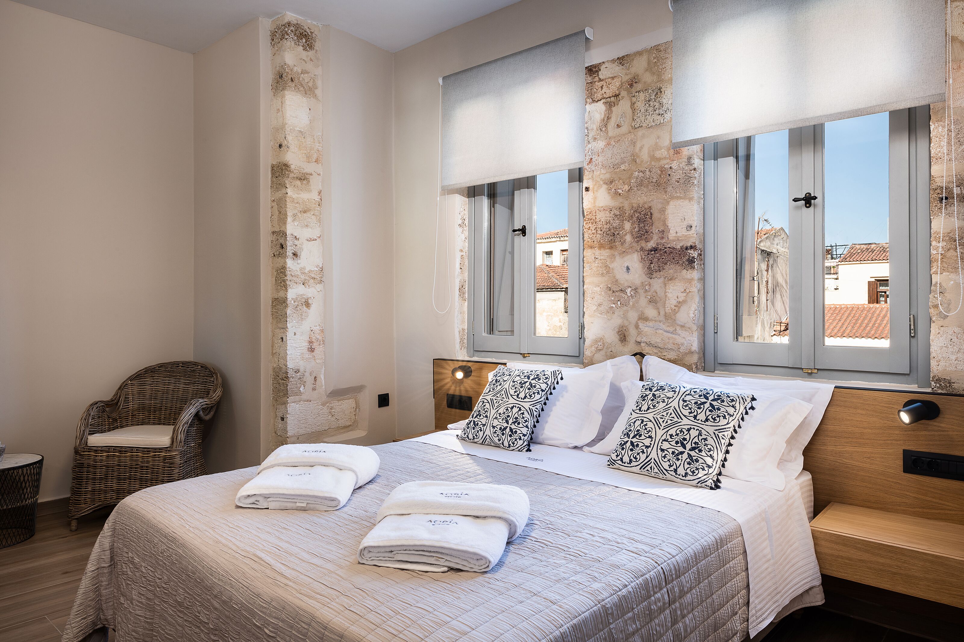 Una camera da letto con un letto, due finestre, una sedia in vimini e vista sugli edifici.