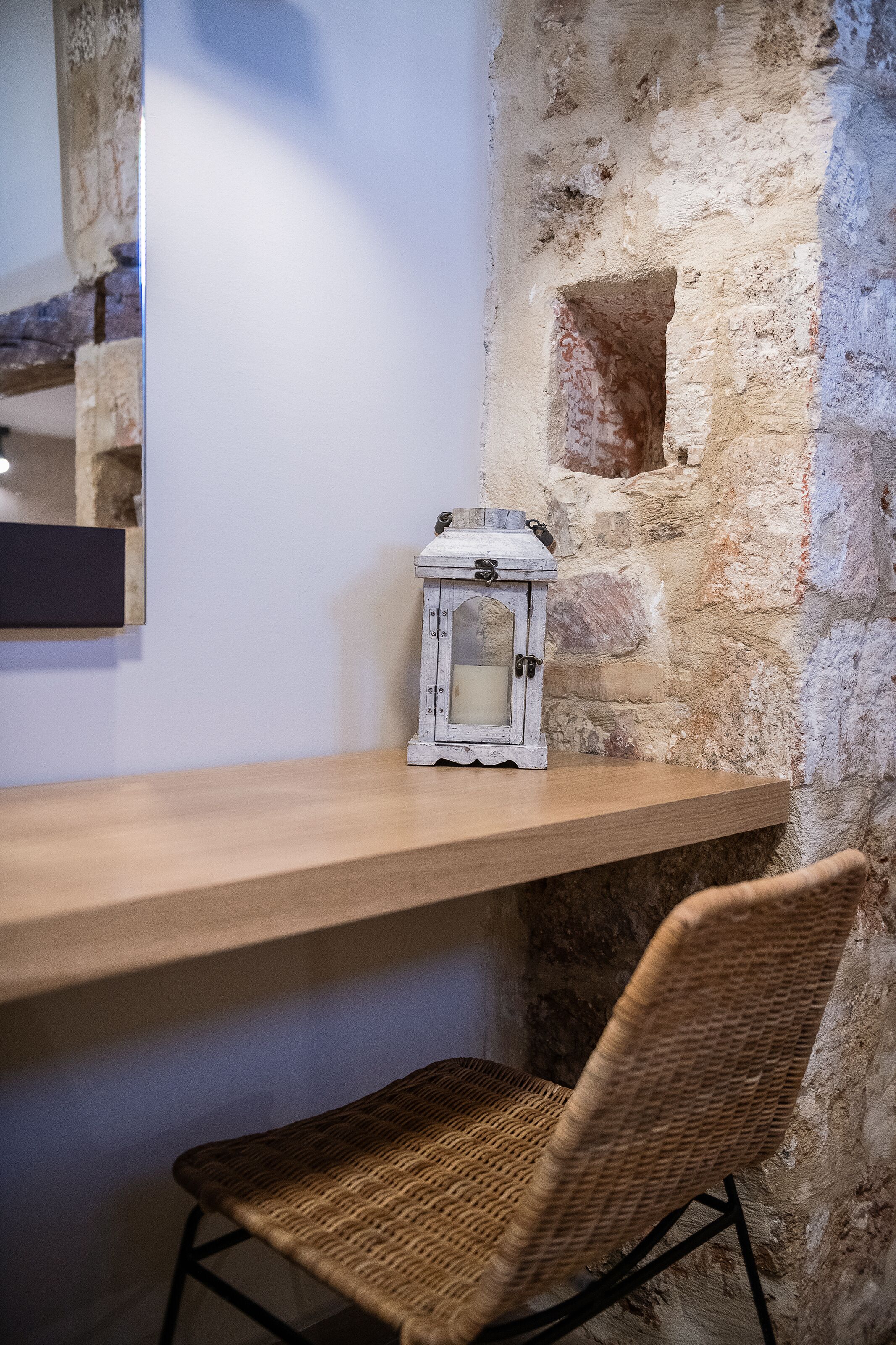 A corner with a wooden shelf, a wicker chair, and a white lantern against a stone wall.