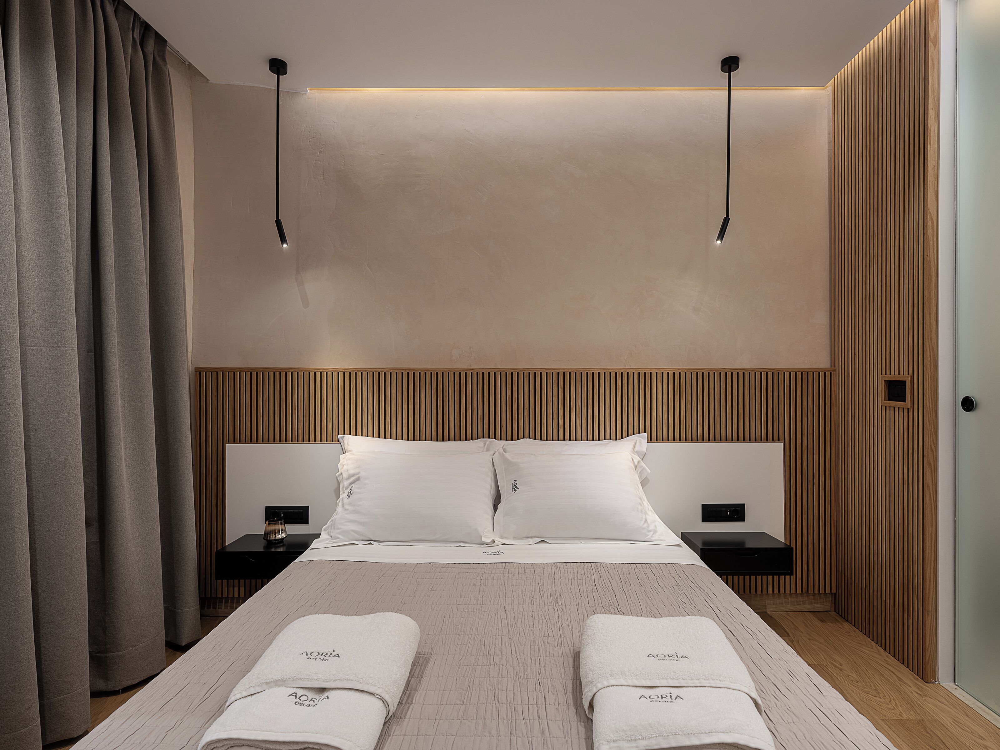 Una habitación de hotel moderna con una cama grande, dos mesitas de noche, un cabecero de madera y un amplio ventanal con cortinas.