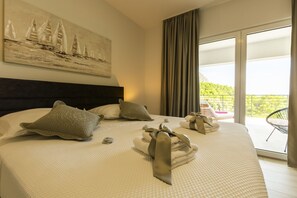 Ein Hotelzimmer mit einem großen Bett, einem Gemälde mit Segelbooten, einem Balkon mit einem Stuhl und Blick auf Grünflächen.
