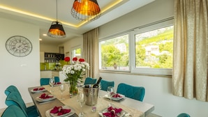 Una sala da pranzo con sedie color petrolio, un tavolo apparecchiato per sei persone, una sveglia appesa al muro e vista sul verde all'esterno.