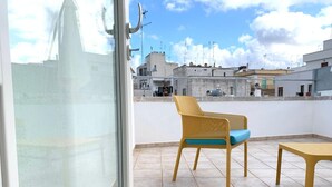 A balcony with a wicker chair and a small table, overlooking a cityscape with buildings and a clear sky. - UNICO - Art & Decor (Ostuni)