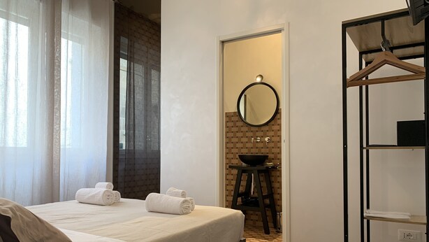 A hotel room with a bed, a bathroom with a sink and mirror, and a wooden storage unit. - UNICO - Art & Decor (Ostuni)