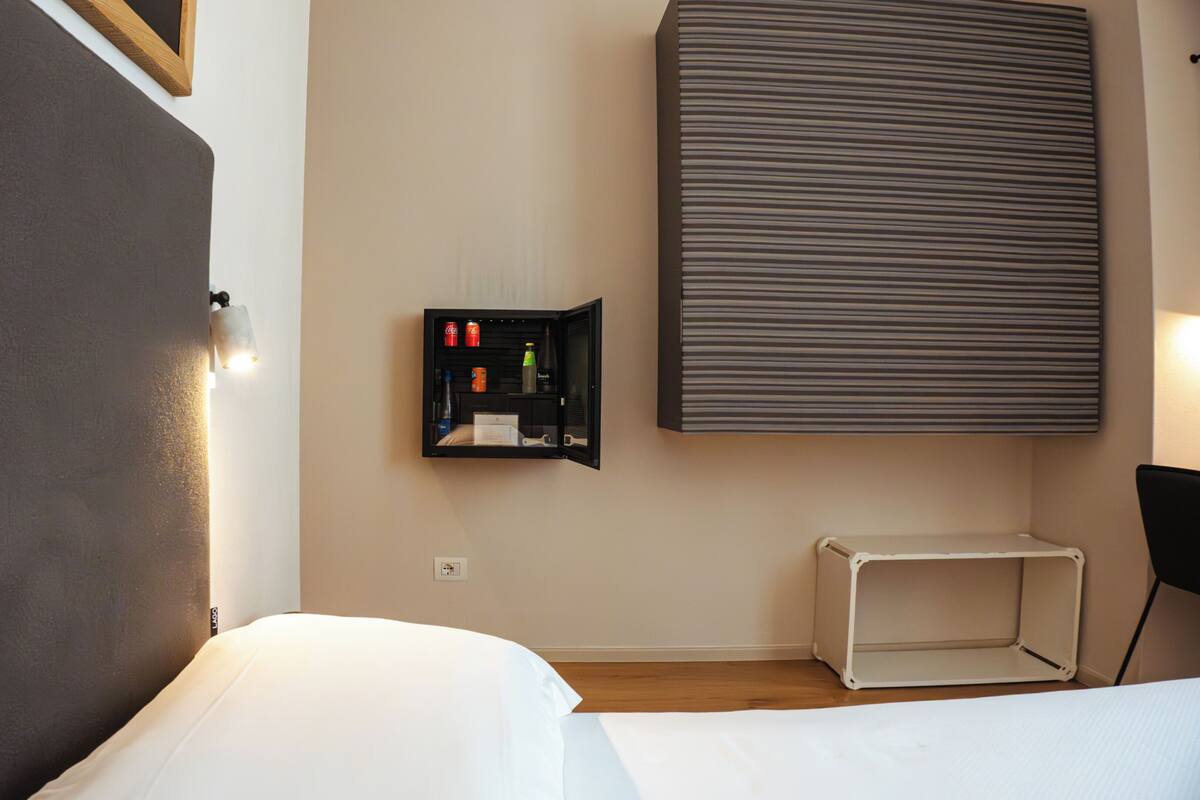A hotel room with a bed, a small cabinet with toiletries, a wall-mounted radiator, and a plain wall.